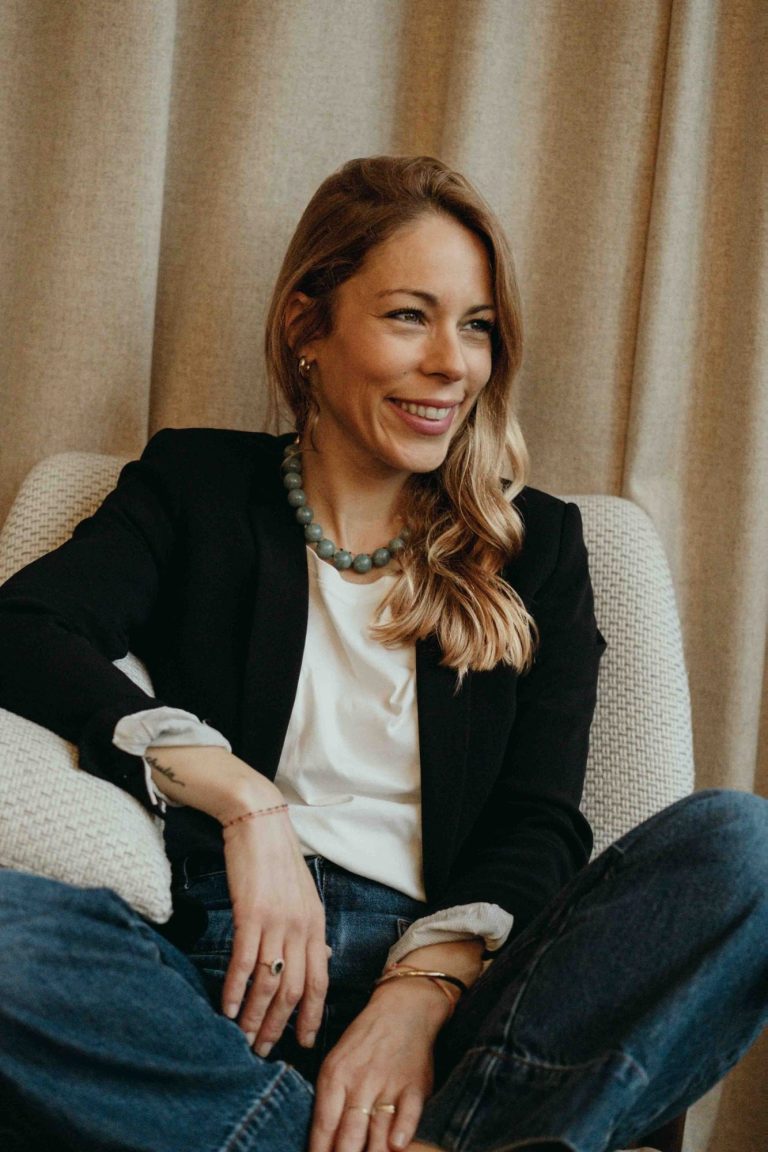 Respira Coaching – Kelly Bisson | Coach développement personnel femmes Bordeaux Femme souriante avec des cheveux blonds, portant un blazer noir et assise sur un canapé.